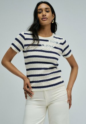 Mujer con cabello largo y oscuro, vestida con un top de punto a rayas blanco y azul marino de manga corta con bordado floral y pantalones blancos, posando con las manos en las caderas.
