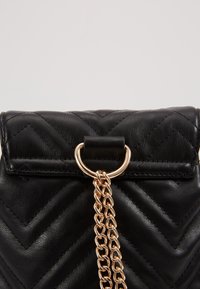 Schwarze gesteppte Lederhandtasche mit umklappbarem Deckel, goldfarbener Kettenverzierung und einem runden Hardware-Ring. Strukturierter Zickzack-Muster auf der Oberfläche.