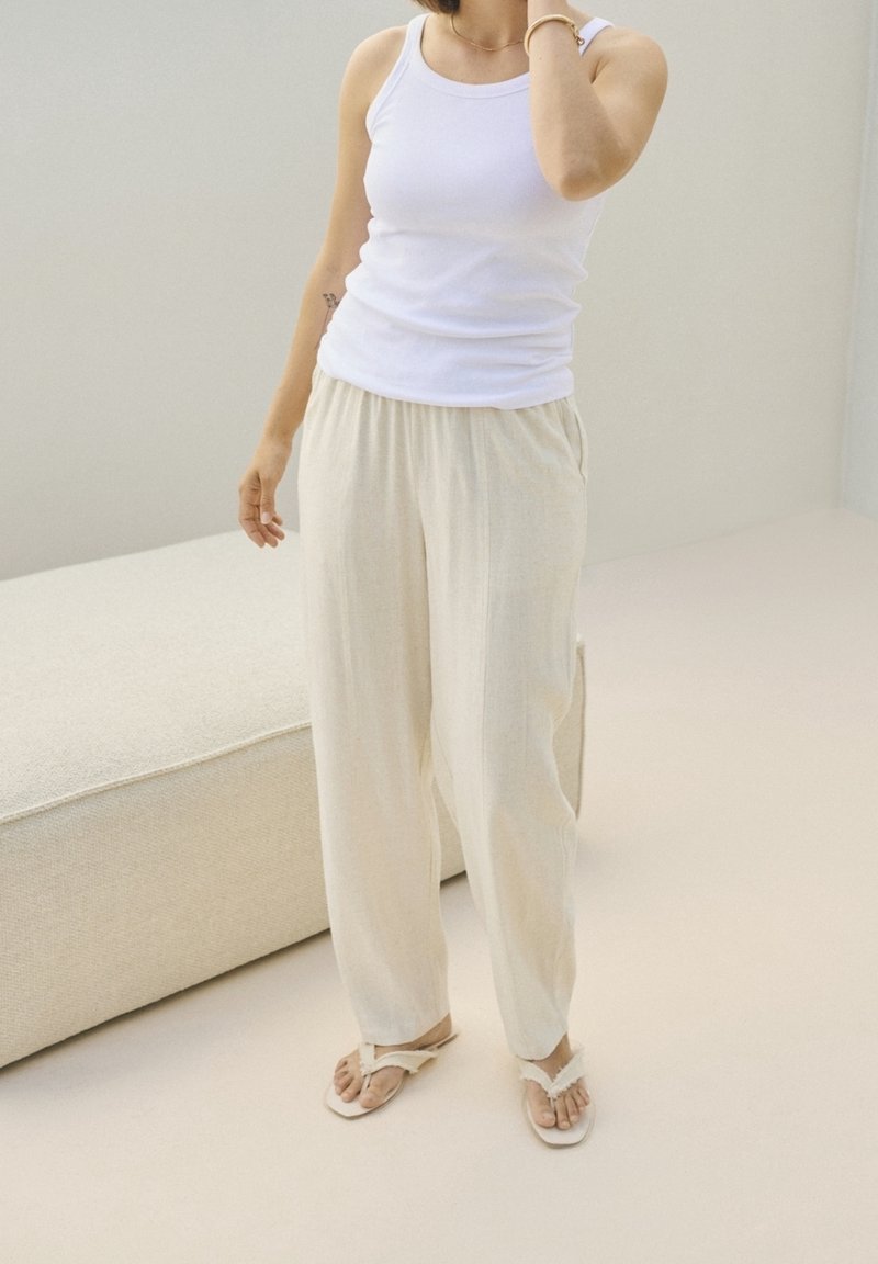 Femme debout à l'intérieur portant un débardeur blanc ajusté, un pantalon ample couleur crème, des sandales blanches et des bijoux légers.