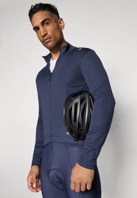 Mann in marineblauem Radoutfit, der einen schwarzen Fahrradhelm vor einem schlichten, hellen Hintergrund hält.