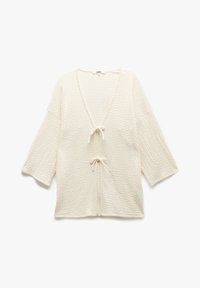 Haut tricot crème avec un col en V, des manches trois-quarts et des liens à l'avant. Le tissu texturé ajoute de la dimension au design.
