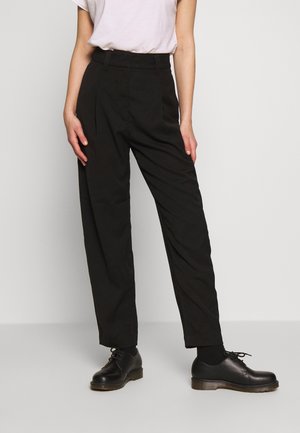 Person trägt hoch taillierte schwarze plissierte Hose, weißes eingestecktes Hemd, schwarze Socken und schwarze Schnürschuhe, steht vor einfachem Hintergrund.