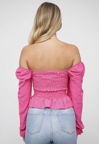 Strak roze off-shoulder top met een geplooide lijfje, lange mouwen en een gerimpelde zoom. Draagt bij lichte blauwe spijkerbroek.