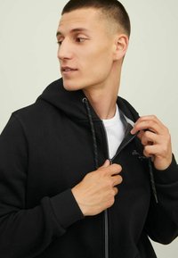 Schwarze Kapuzenjacke mit Reißverschluss, Kordelzug und bestickten Details. Weicher Stoff, taillierte Passform, sichtbarer Reißverschluss und kontrastierendes Innenfutter.