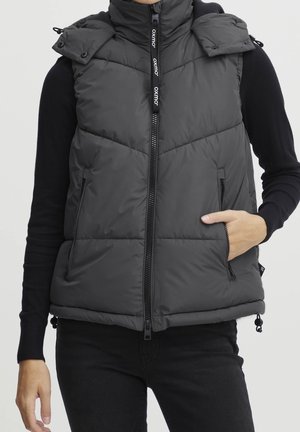 Schwarze Steppweste mit Frontreißverschluss und zwei Reißverschlusstaschen, getragen über einem schwarzen Langarmshirt von einer Person mit einer Hand in der Tasche.