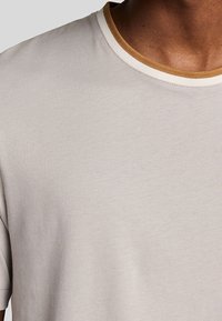 Camiseta gris claro con cuello redondo. El cuello cuenta con un ribete acanalado en crema y mostaza en contraste. Textura suave, manga corta.