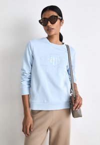 Világoskék pulóver fehér, kiemelkedő hímzéssel, kerek nyakkal és bordázott mandzsettával. Barna nadrággal és egy kis szürke táskával párosítva.