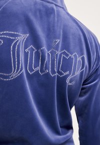 Kék bársony kapucnis pulóver, melynek hátán „Juicy” felirat csillogó gyémántdíszítéssel, sima textúrát és kényelmes kialakítást kínál.