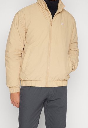 Mann trägt eine beige Reißverschlussjacke über einem schwarzen Rollkragenpullover und dunkelgrauen Hosen, mit einer Hand in der Jackentasche.
