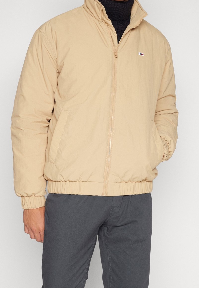 Homme portant une veste beige zippée sur un col roulé noir et un pantalon gris foncé, avec une main dans la poche de la veste.