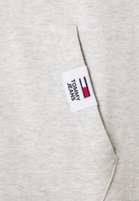 Tissu gris clair avec une poche avant dotée d'une étiquette blanche mentionnant "TOMMY JEANS" et un logo horizontal rouge, blanc et marine.