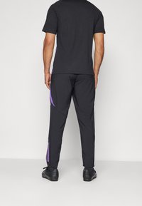 Zwarte kortemouwen shirt gecombineerd met zwarte broek met paarse zijaccenten. De broek heeft ritsen bij de enkels en een gladde textuur.