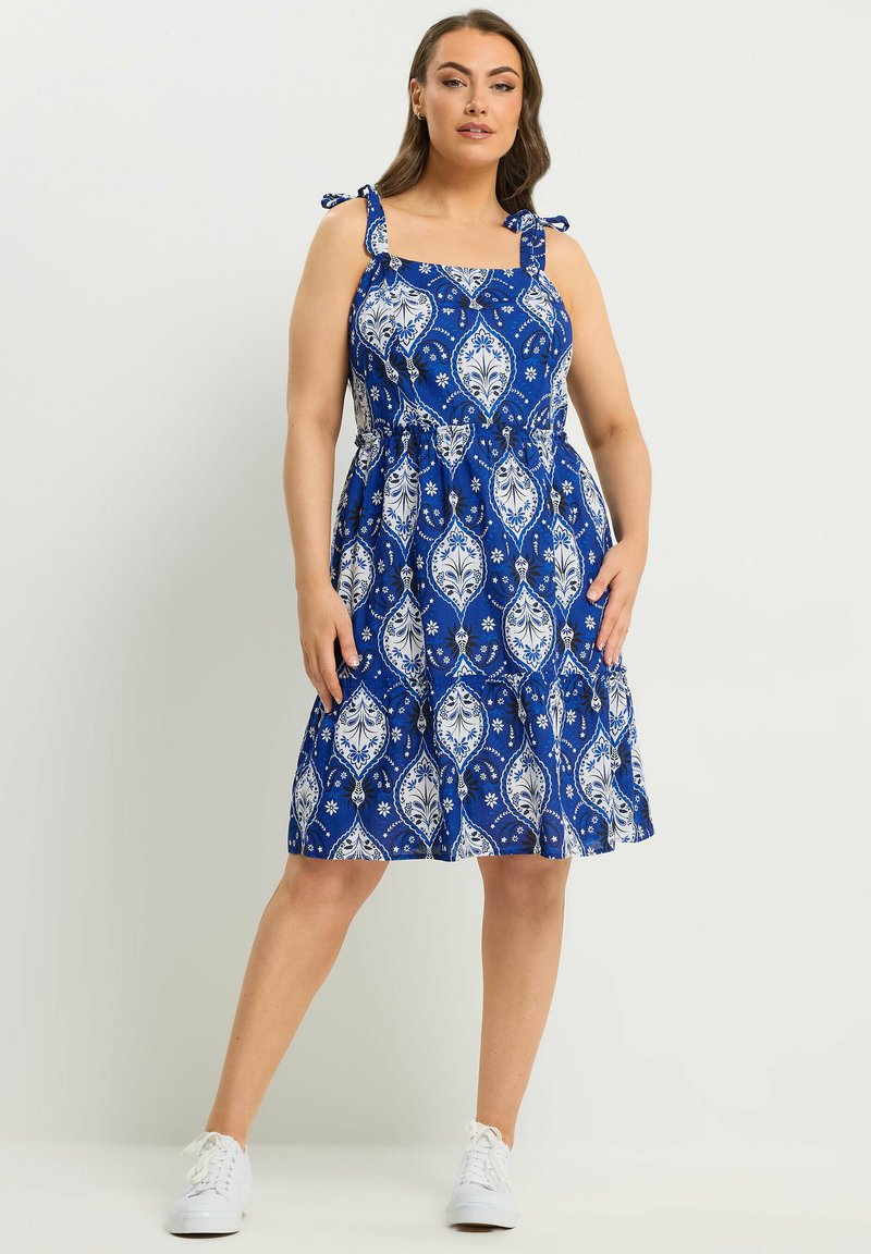Frau in knielangem blauem Kleid mit weißen und marineblauen Blumenmustern, gebundenen Schulterträgern und weißen Turnschuhen, die mit einem Bein nach vorne steht.