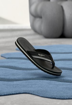 Černé žabky s bílým pruhem a logem "Pepe Jeans" plují nad modrým texturovaným kobercem, na pozadí je velký rozmazaný šedý polštář.