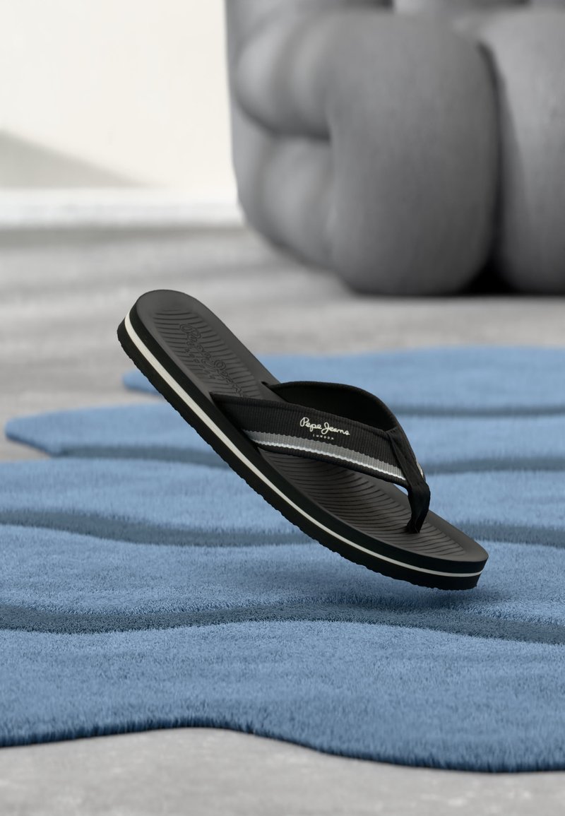 Μαύρη σαγιονάρα με λευκή ρίγα και λογότυπο "Pepe Jeans" αιωρείται πάνω από μπλε υφασμάτινο χαλί, με ένα μεγάλο θολό γκρι μαξιλάρι στο φόντο.