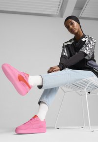 Sneaker in suede rosa con dettagli arancioni, caratterizzati da un design low-top, una parte superiore testurizzata e suola piatta. Indossati con jeans azzurri e calzini bianchi.