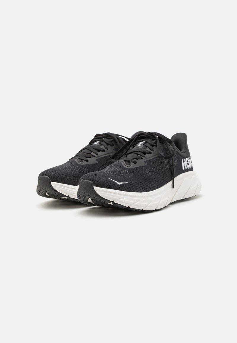 Scarpe sportive nere con tomaia in rete testurizzata, suola intermedia bianca e logo prominente. Punta arrotondata, lacci piatti e suola esterna in gomma con battistrada.