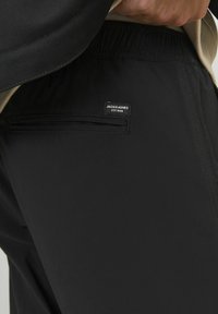 Sorte bukser med elastisk talje, der har en lynlås lomme og et mærke med "JACK & JONES EST. 1989" bagpå. Glat, mat stofstruktur.