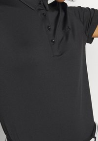 Personne portant un polo à manches courtes noir avec des boutons et un petit logo Under Armour foncé sur la poitrine.