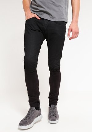 Homme portant un pantalon noir slim, un t-shirt gris et des baskets en daim gris, debout avec une main dans la poche.