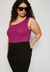 Top de tirantes morado ajustado con textura acanalada, combinado con pantalones negros con bolsillos. La modelo lleva gafas de sol oscuras, mostrando una vista lateral.