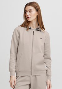 Svetlo béžová mikina na zips s nastaviteľnými čiernymi šnúrkami, klokankovými vreckami a diskrétnym logom na hrudi. Mäkko hladký materiál.