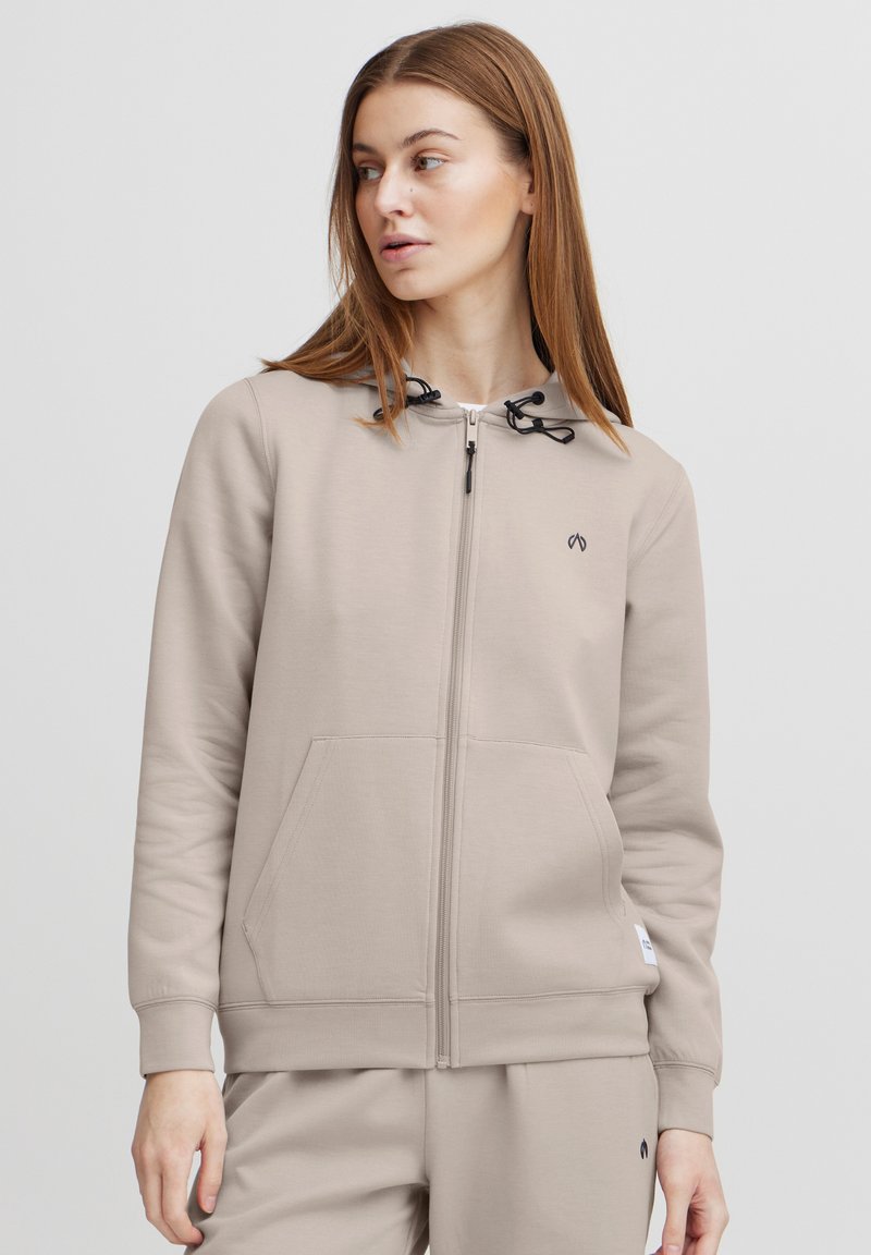 Svetlo béžová mikina na zips s nastaviteľnými čiernymi šnúrkami, klokankovými vreckami a diskrétnym logom na hrudi. Mäkko hladký materiál.