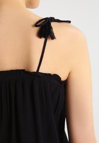 Top senza maniche nero con scollo arricciato, spalline sottili e decorazioni a nappina sulle spalle, realizzato in un tessuto leggero.