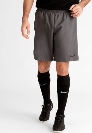 Homme portant un short de sport gris Nike, des chaussettes de football noires et des crampons noirs, debout avec jambes et mains visibles sur fond blanc.