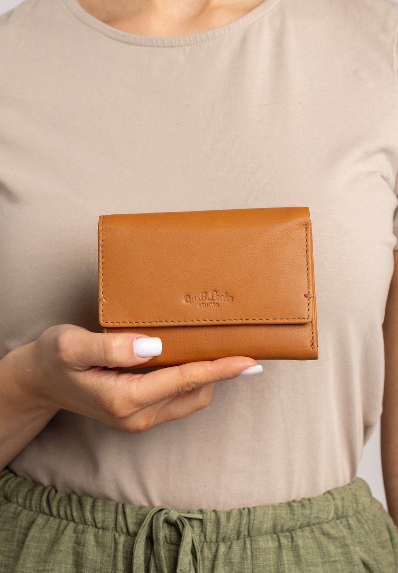 Brązowy skórzany portfel o eleganckim designie, z zapięciem na zakładkę i wytłoczonym logo. Charakteryzuje się starannym przeszyciem i gładką teksturą.