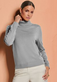 Grau meliertes Rollkragenpullover mit gerippten Bündchen und Saum, aus weichem Material, kombiniert mit hellen Hosen vor einem pfirsichfarbenen Hintergrund.