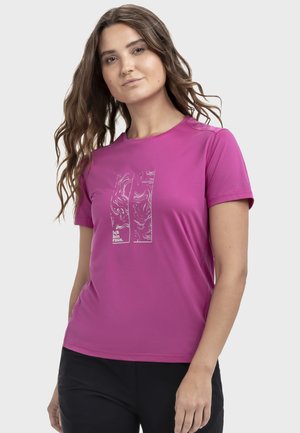 Woman with wavy brown hair wearing a magenta short-sleeve shirt with abstract white design and "Ich bin raus." text, paired with black pants.