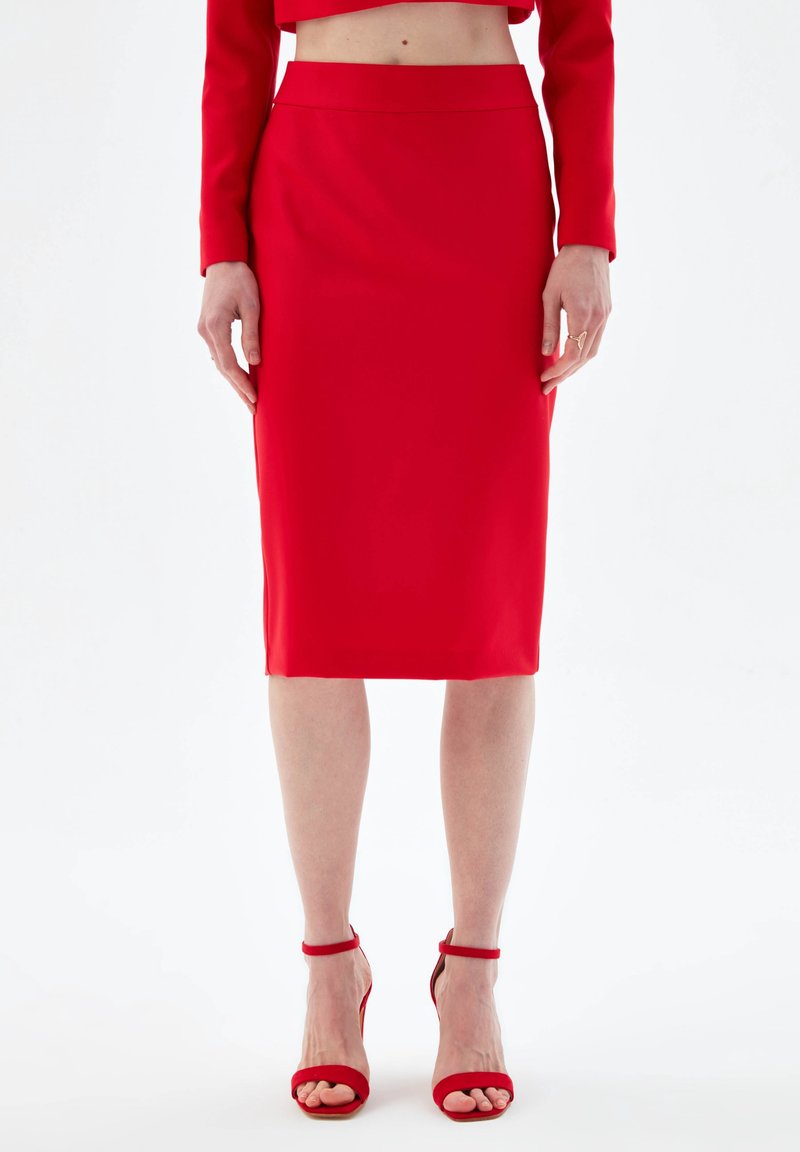 Femme portant une jupe crayon rouge taille haute et des sandales à talons aiguilles rouges assorties avec bride cheville, debout sur un fond blanc.