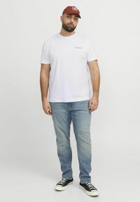 Weißes Baumwoll-T-Shirt mit einem auf der linken Brust aufgedruckten Logo, kombiniert mit hellblauen Jeans und schwarzen Leinenschuhen. Rote Mütze getragen.