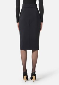 Jupe crayon noire à taille haute, fermeture éclair, et deux poches avant, associée à des chaussures à talons hauts noires et des collants noirs transparents.