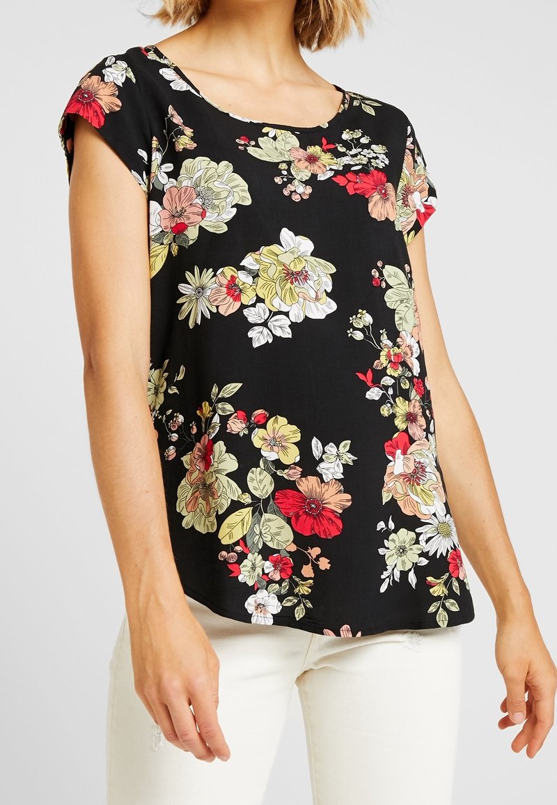 Femme portant un chemisier noir à manches courtes avec un imprimé floral coloré et un pantalon blanc, debout devant un fond uni.