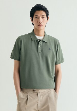 Hombre joven con cabello oscuro, vestido con una camisa tipo polo verde y pantalones beige, de pie con las manos en los bolsillos contra un fondo liso.
