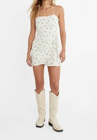 Weißer floral gemusterter Jumpsuit mit quadratischem Ausschnitt und dünnen Trägern, ausgestattet mit einer seitlichen Schleife. Kombiniert mit cremefarbenen Cowboy-Stiefeln mit aufwendigem Design.