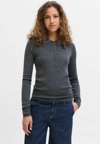 Žena s kudrnatými vlasy v tmavě šedém dlouhém polo svetru a modrých džínách, stojící na jednobarevném bílém pozadí.