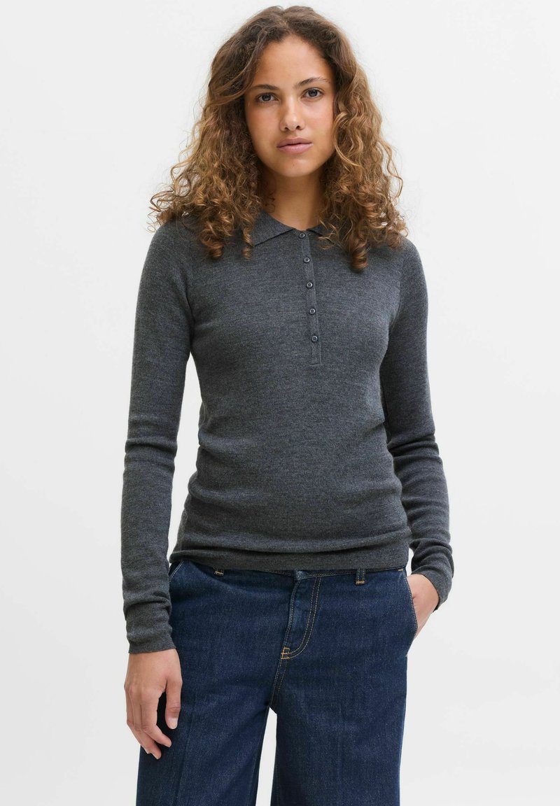 Žena s kudrnatými vlasy v tmavě šedém dlouhém polo svetru a modrých džínách, stojící na jednobarevném bílém pozadí.