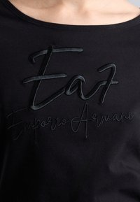 Melna T krekls ar izvirzītu melnu izšuvumu "EA7 Emporio Armani" pār krūtīm, ar gludu, pieguļošu dizainu un apaļu dekoltē.