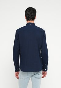 Tamsiai mėlynas ilgomis rankovėmis marškinėlis su sagomis ant apykaklės, matomais siūlais ir perlo formos snapais rankogaliuose bei užpakalinėje dalyje. Lygi tekstūra.