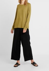 Olive green long-sleeve shirt with round neckline, paired with black wide-leg trousers and black slide sandals. Soft fabric with side slits.