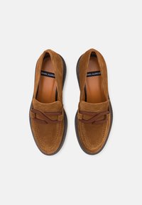 Mocasines de ante marrón con punta redonda, que presentan un diseño bordado y una banda decorativa marrón en la parte frontal. Suelas de goma planas.