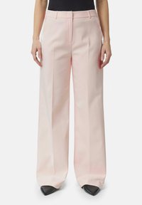 Pantaloni a gamba larga rosa chiaro realizzati in tessuto liscio. Presentano una piega centrale, passanti per cintura e una chiusura frontale con zip. Scarpe nere con punta affusolata.