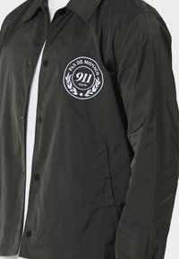 Czarna kurtka zapinana na guziki z białym, okrągłym logo "Pas de Monaco 911 Club" na lewej piersi oraz kieszenią z rozcięciem poniżej.