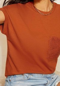 Tričko v barvě rezavé s texturovanou háčkovanou kapsou, krátké rukávy a uvolněný střih, spojené se světle modrými džínovými šortkami.