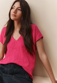 Haut fuchsia à manches courtes avec col en V et tissu texturé, associé à un pantalon en denim foncé taille haute, sur fond neutre.