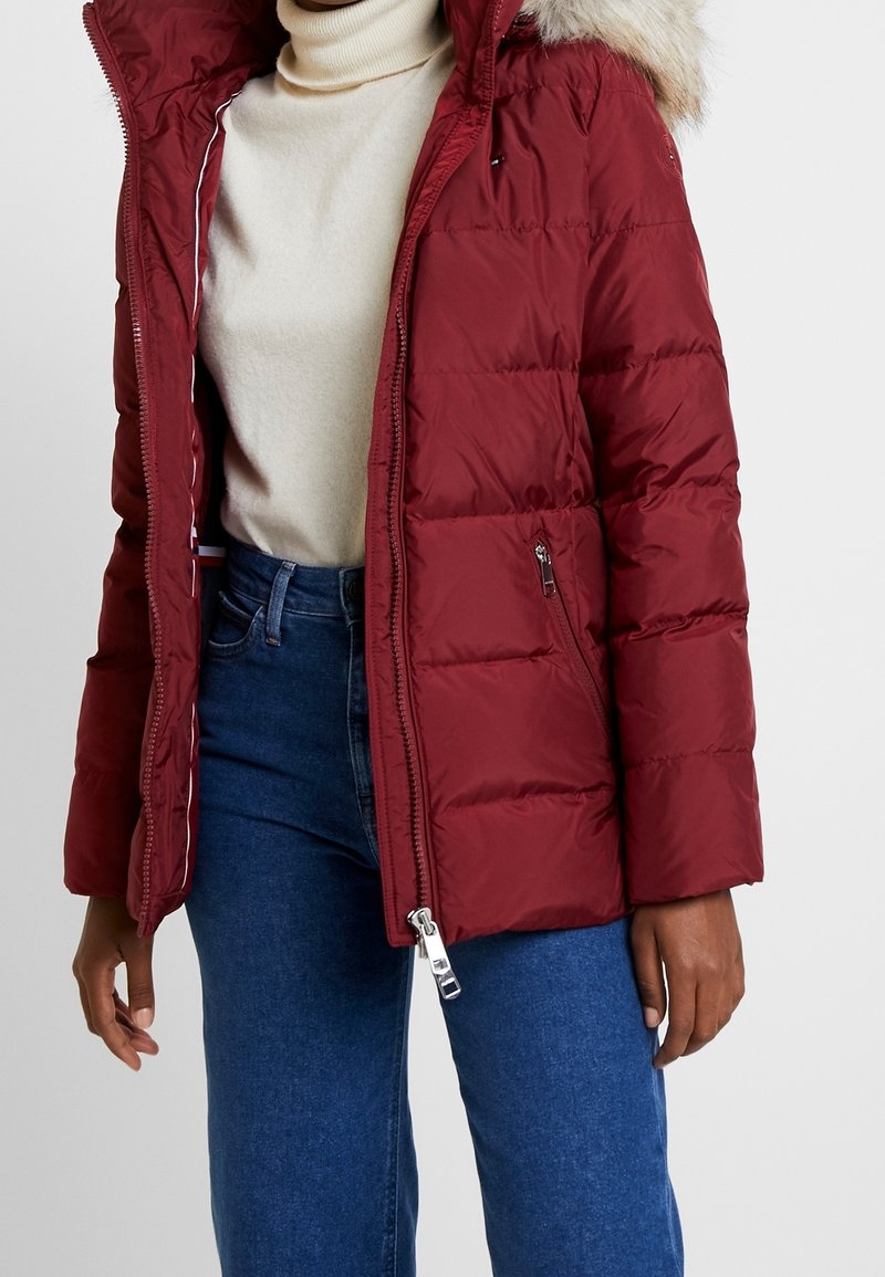 Person som bär en mörkröd vadderad jacka med pälskantad luva, beige polotröja och blå jeans, stående mot en vit bakgrund.