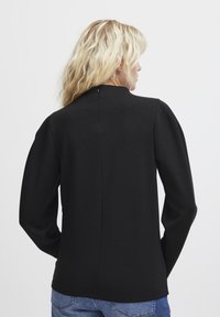 Maglietta nera a maniche lunghe con un collo alto, spalle gonfie e una texture liscia. Il retro presenta un dettaglio di chiusura discreto.
